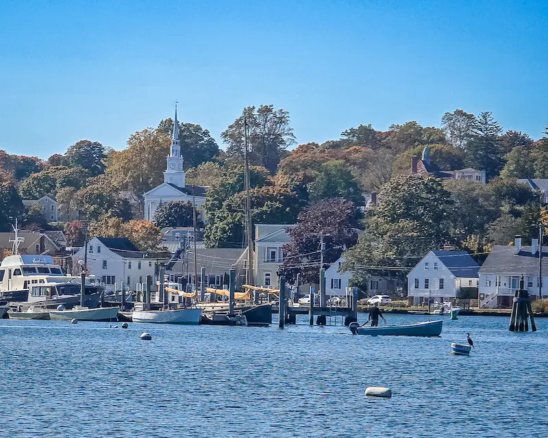 A New England harbor town with a white church steeple above boats and docks at the water’s edge