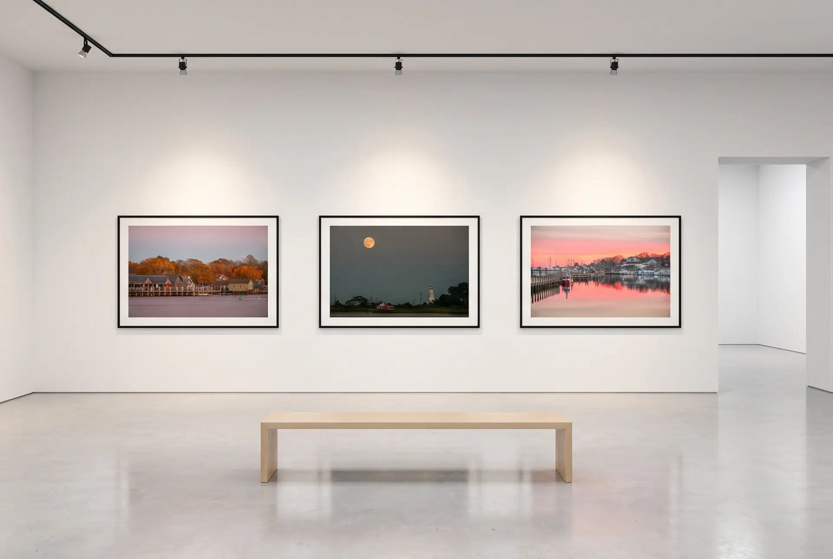 Three framed coastal photographs by Sandra Falcetta on a white gallery wall above a wooden bench